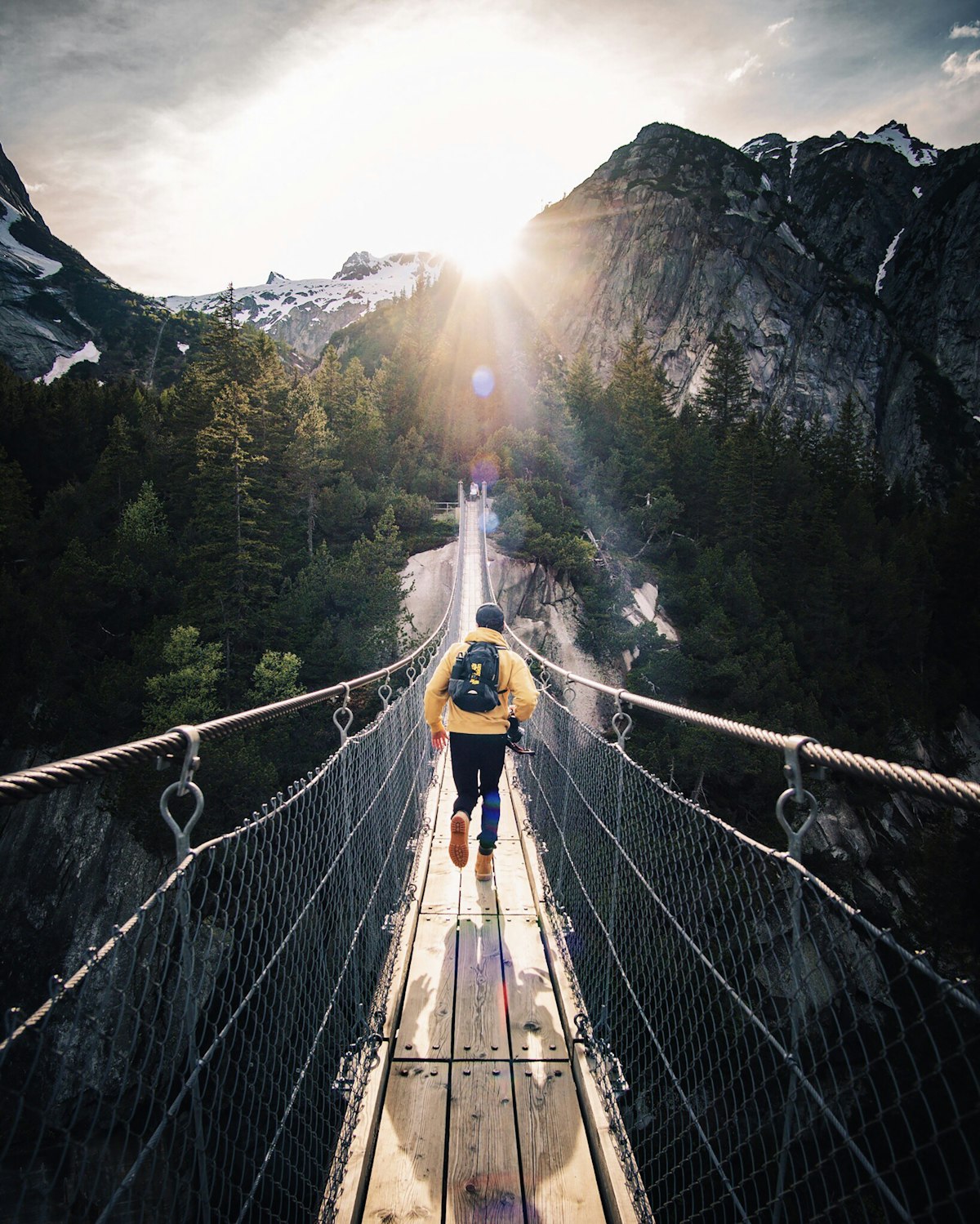 A traveler crossing a suspension bridge toward snow-capped peaks